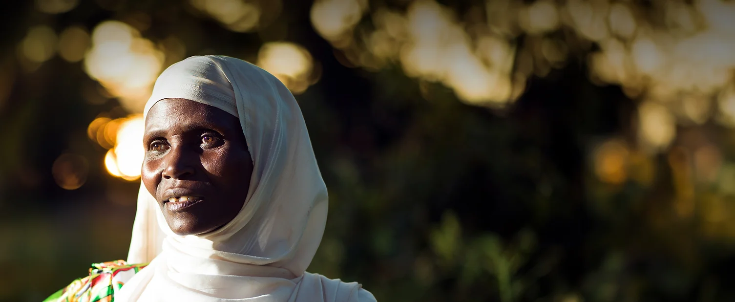 Woman in white headscarf smiling outdoors with sunlight behind.