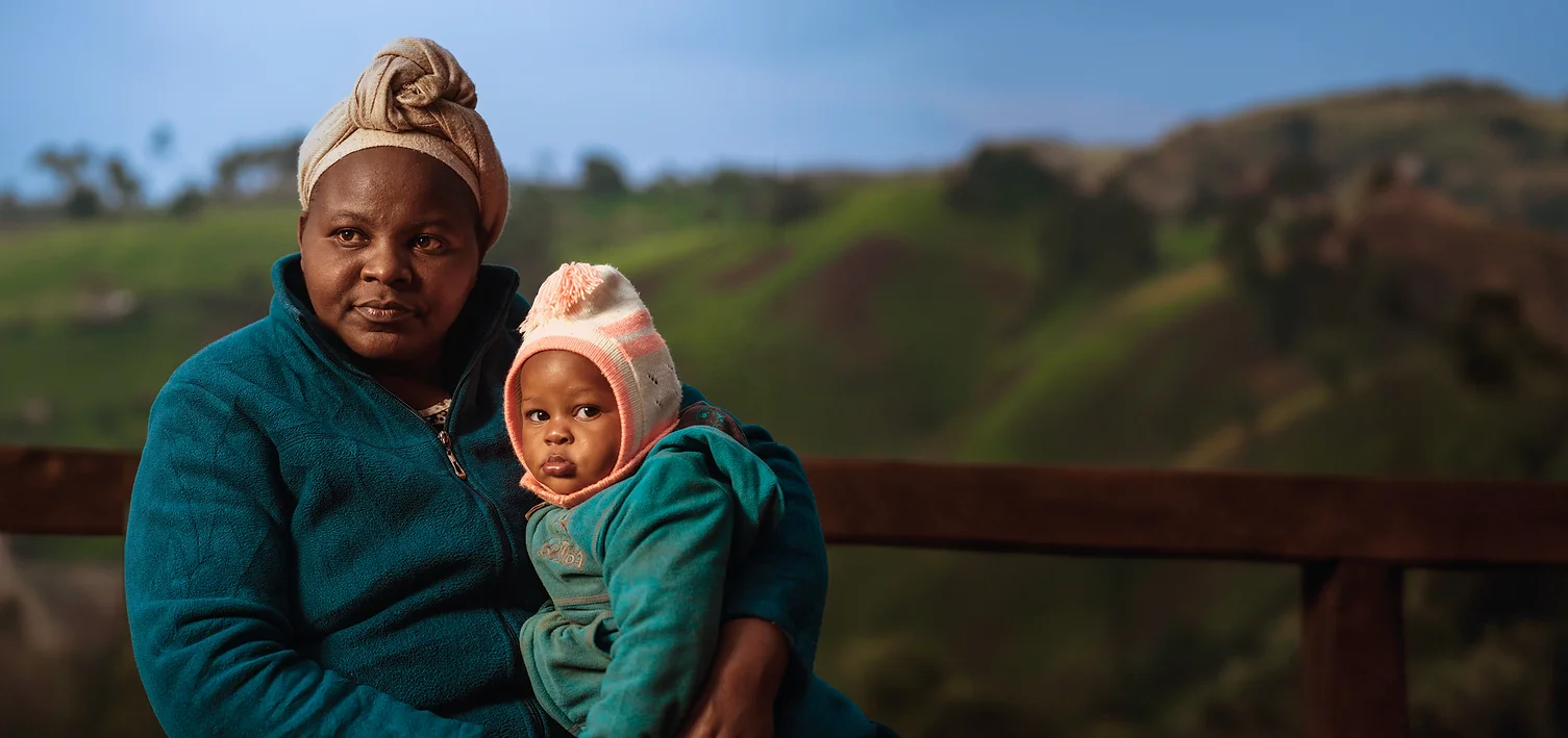 Woman with child in blue jackets against a scenic hillside.