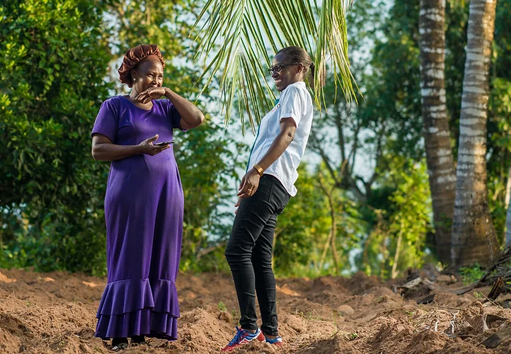 Two women laughing and talking under a palm tree outdoors.